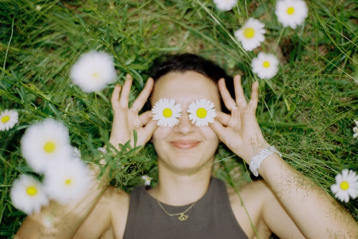 Film photograph of a person lying in a meadow holding two daisies over their eyes