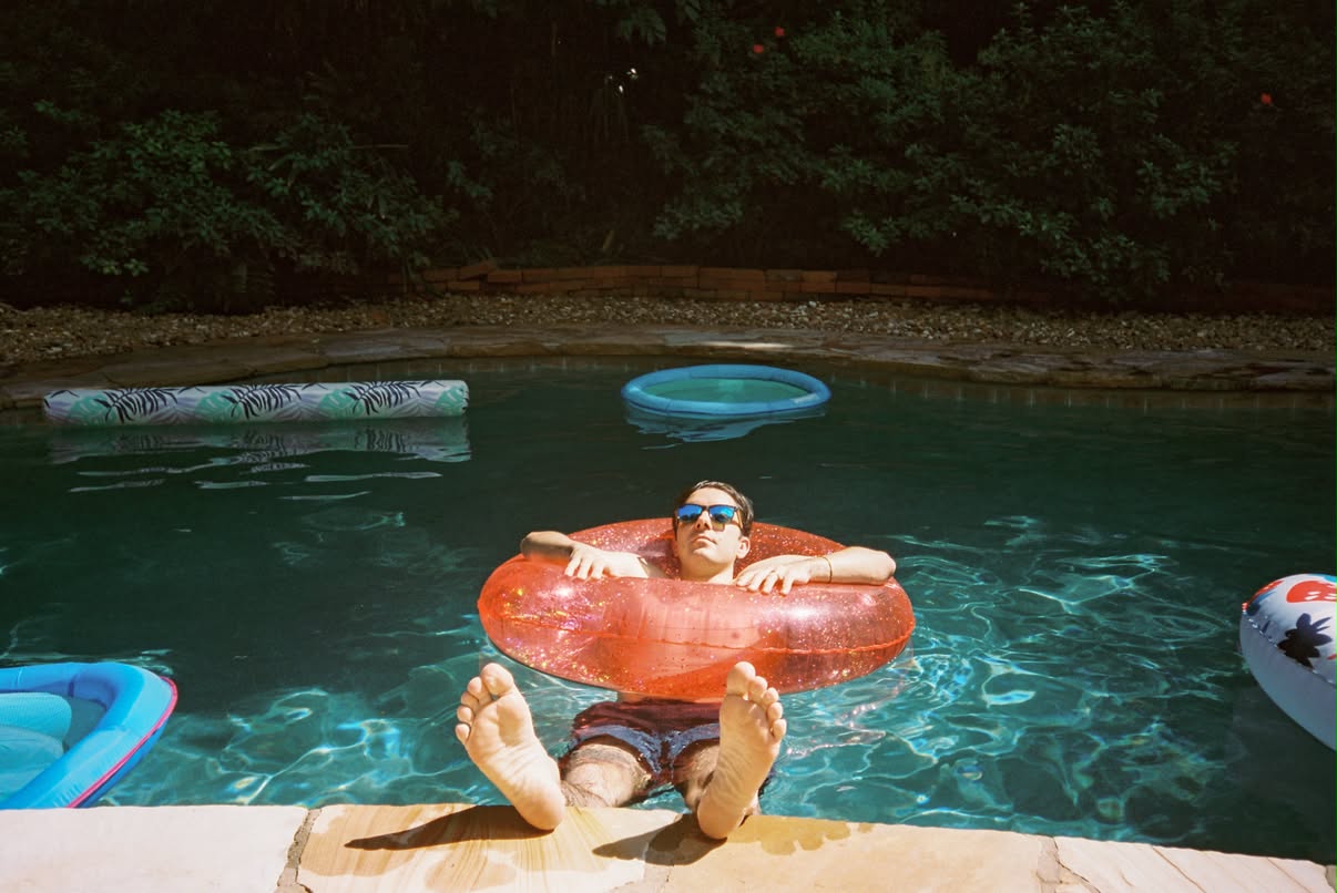Film photograph of a man in sunglasses relaxing in a red glitter inner tube in a green swimming pool