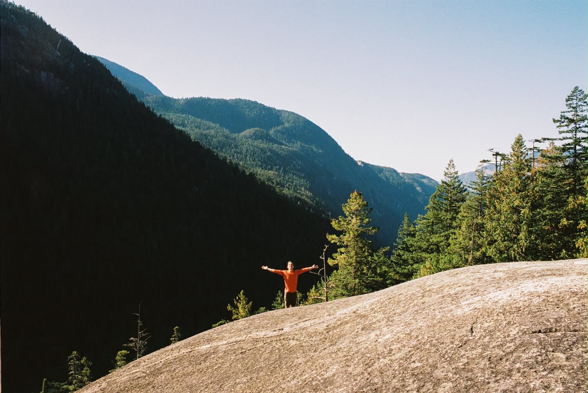 Film photograph of a person with arms outstretched atop a granite dome overlooking forested mountains near Squamish, Canada