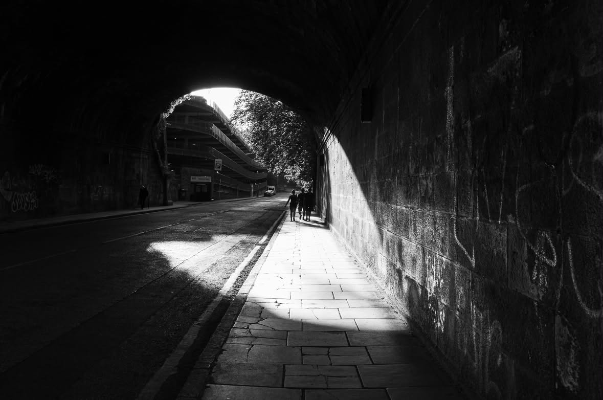 Black-and-white photograph looking through a stone tunnel toward light, with small figures walking in the distance