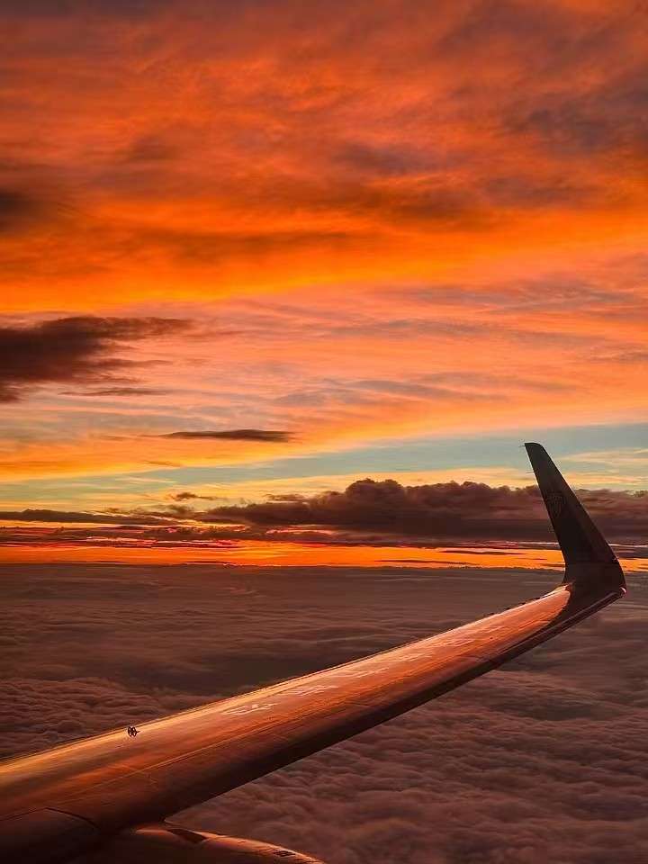 Photograph of an airplane wing silhouetted against a fiery orange sunset above a sea of clouds