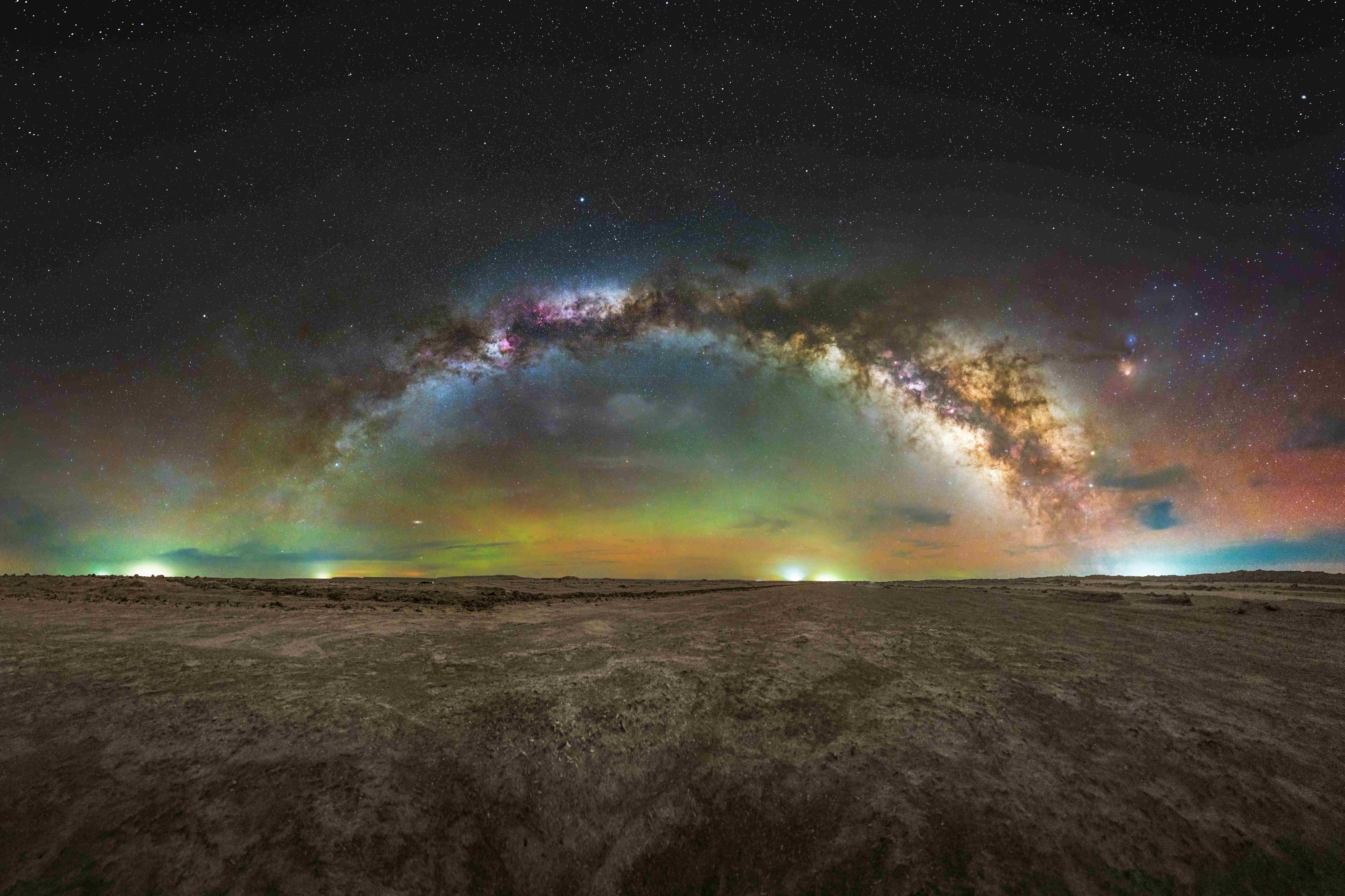 Photograph of the Milky Way arcing over a dark desert landscape, with green and orange aurora-like glow along the horizon