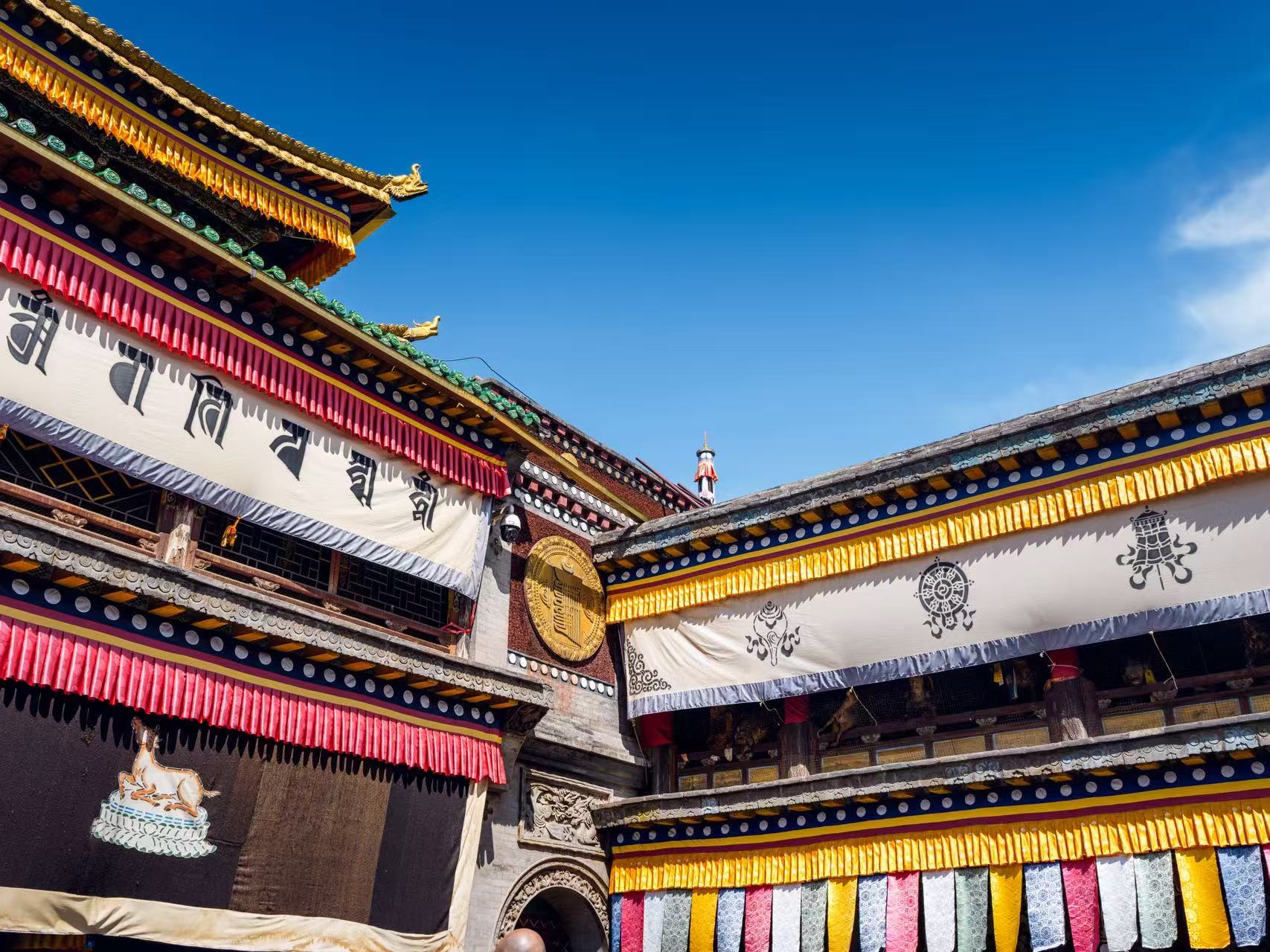 Photograph of colorful patterned banners and golden roof details on a Tibetan monastery against a clear blue sky