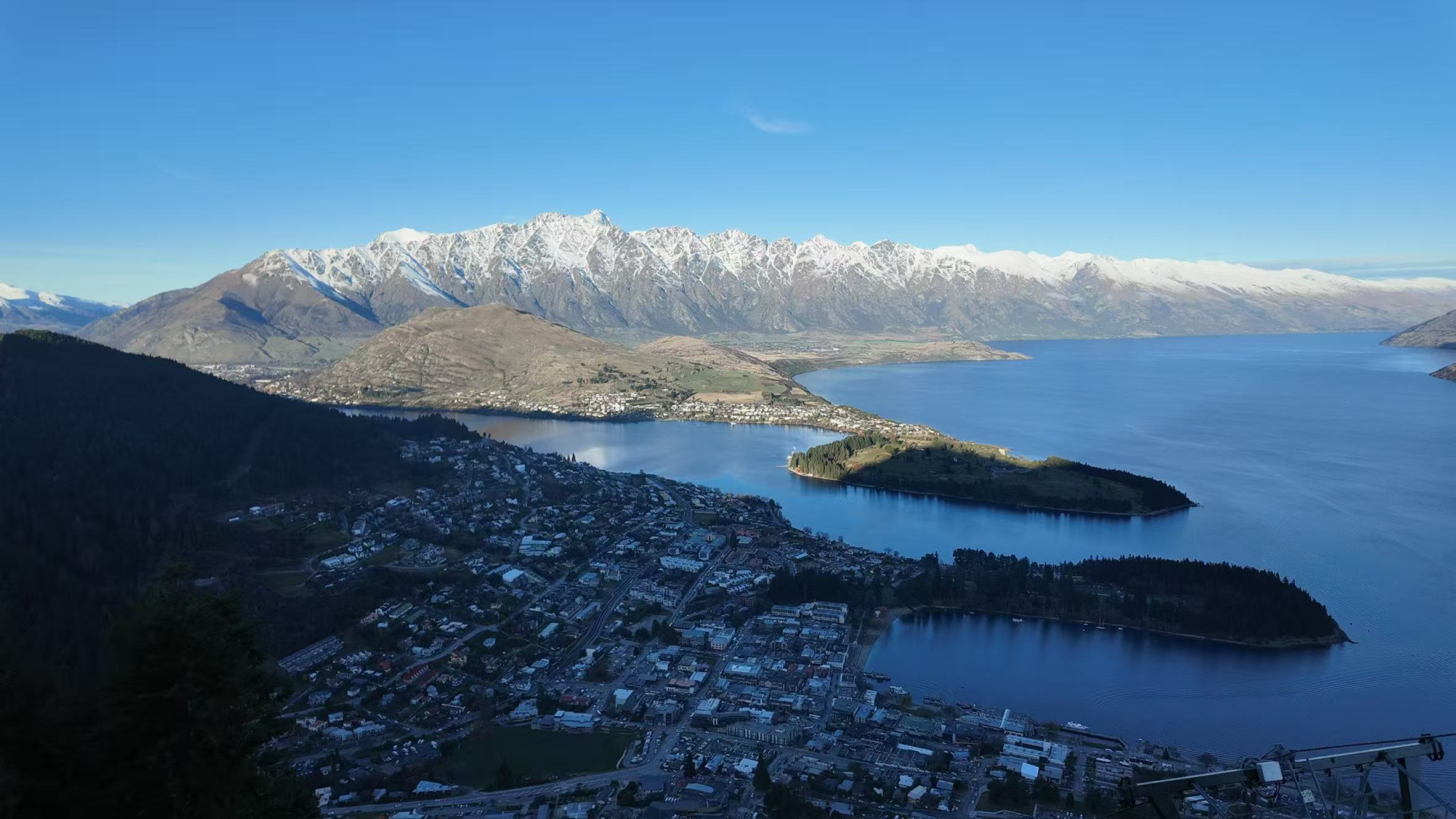 Aerial photograph of Queenstown, New Zealand, with a blue lake, forested peninsula and the snow-capped Remarkables mountain range behind