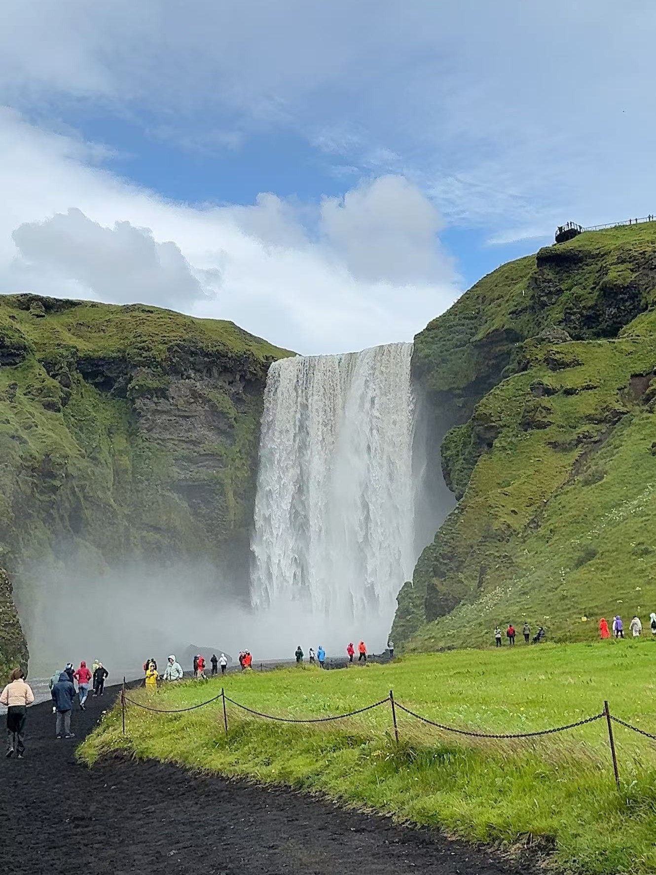 Photograph of Skogafoss waterfall in Iceland cascading between green cliffs, with visitors in colorful raincoats gathered below