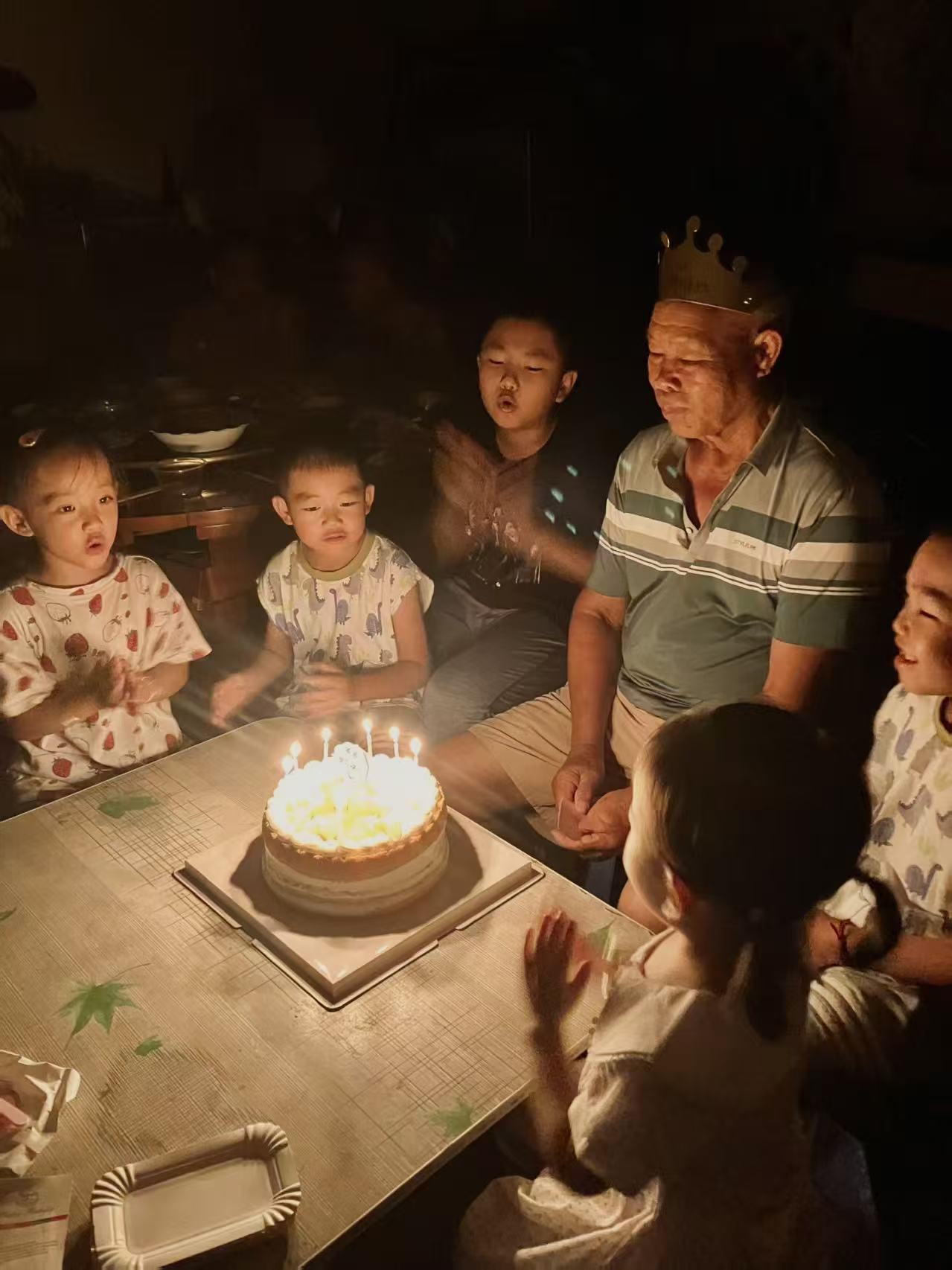 Photograph of a grandfather wearing a paper crown surrounded by his grandchildren around a candlelit birthday cake