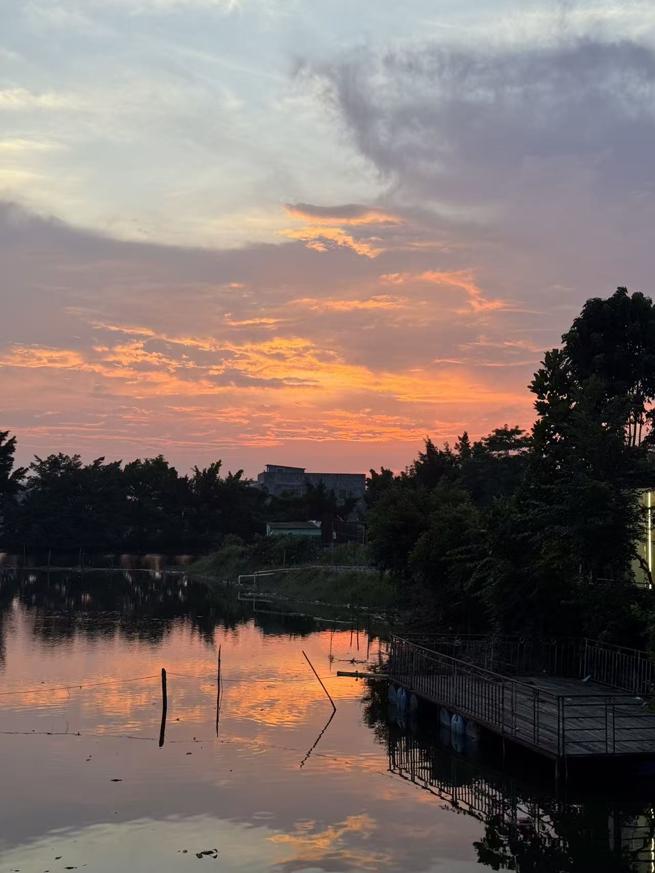 Photograph of a dramatic orange and pink sunset sky reflected in a still lake, with wooden poles and a dock in the foreground